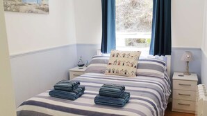 A neatly made bed with striped bedding and a pillow with a barn pattern, two folded towels on the bed, a bedside table with a lamp, and a window with blue curtains. - Foxes Lair  Holiday Home Looe Cornwall (Looe)