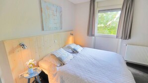 A bedroom with a bed, a small table with a vase of flowers, a wall-mounted light, and a window with curtains. - Duinland 49a Holiday home Sint Maartenszee (Sint Maartensvlotbrug)