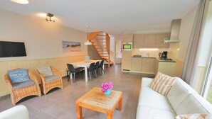 A modern living room with a sofa, two wicker chairs, a coffee table, and a dining area with a table and chairs. A staircase leads to an upper level. - Duinland 49a Holiday home Sint Maartenszee (Sint Maartensvlotbrug)