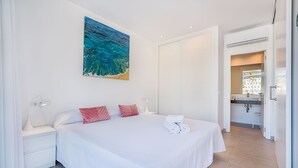 A bedroom with a large bed, white bedding, a painting of a blue ocean, a white nightstand, and a bathroom visible through an open door. - MIRADOR BLUE G   Ref: CS20 (Cala Sant Vicenç)