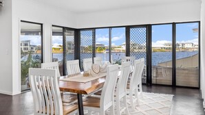 A dining area with a wooden table and white chairs, large windows with a view of the sea and buildings, and a white floor.