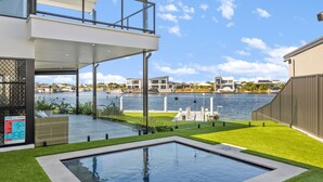 A modern outdoor pool area with a view of the water and boats, a glass-enclosed deck, and a grassy lawn.