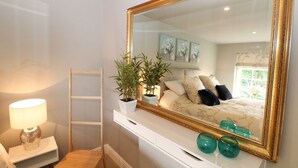 Un dormitorio con un espejo grande, una mesita de noche con lámpara, una silla de madera, una planta en maceta y jarrones de vidrio decorativos.