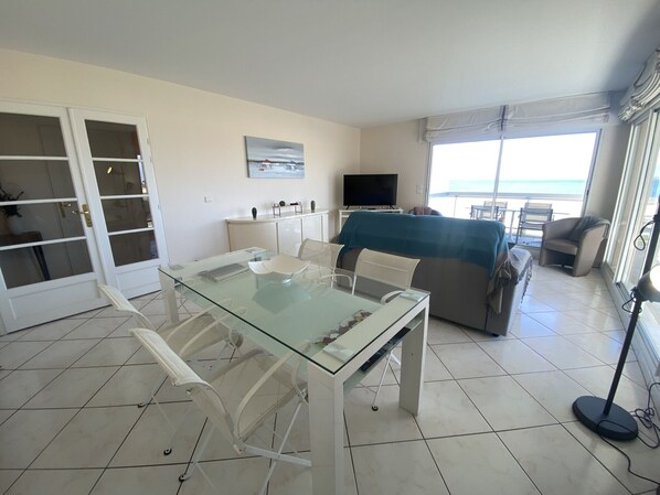 A modern living room with a glass dining table, white chairs, a sofa, and a TV.