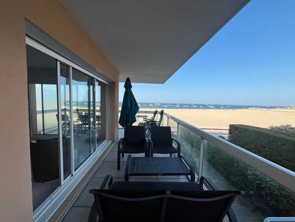 A balcony with a view of the beach, a table, chairs, and a green umbrella.