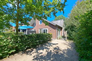 Une maison en briques avec une allée en gravier, entourée d’une végétation luxuriante et d’arbres.