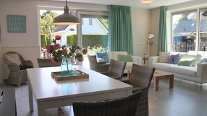 Un soggiorno moderno con un divano bianco, poltrone in vimini, un tavolo da pranzo con un vaso di fiori e ampie finestre con tende.