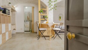 Uma área de refeições com uma mesa e cadeiras de madeira, uma cozinha ao fundo e uma parede de pedra com plantas penduradas.