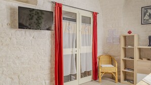 Um quarto com parede de tijolos brancos, uma porta de correr com cortinas vermelhas, uma cadeira de vime, uma estante de madeira e uma televisão de tela plana fixada na parede.