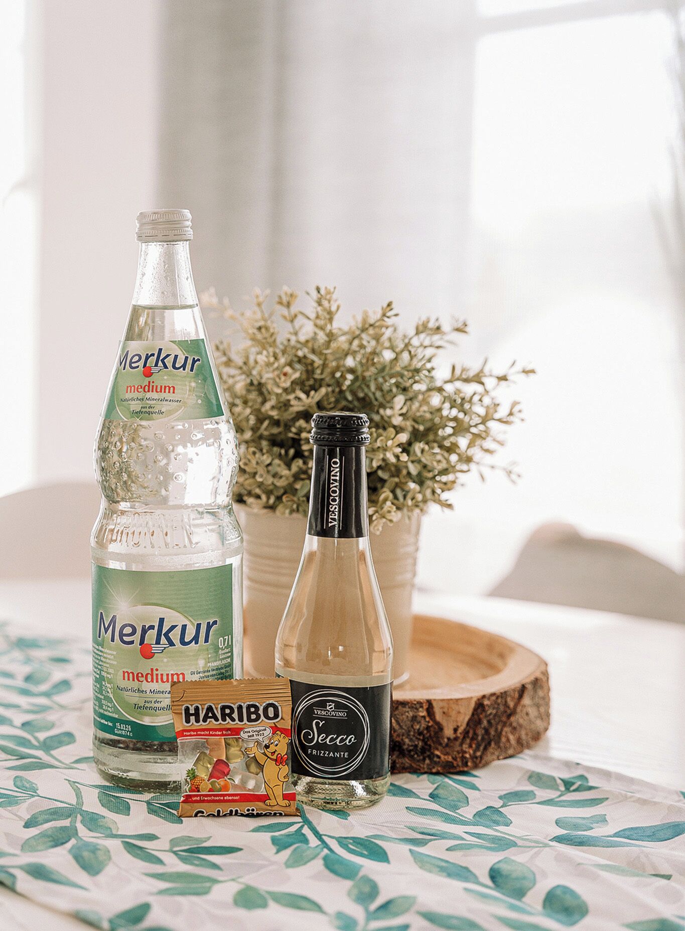 Una botella de agua mineral Merkur mediana, una botella de Secco Frizzante y una barra de caramelos Haribo sobre una mesa con mantel floreado.