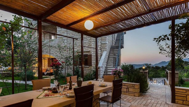 An outdoor dining area with a stone wall backdrop, a wooden pergola, and a view of a mountain range.