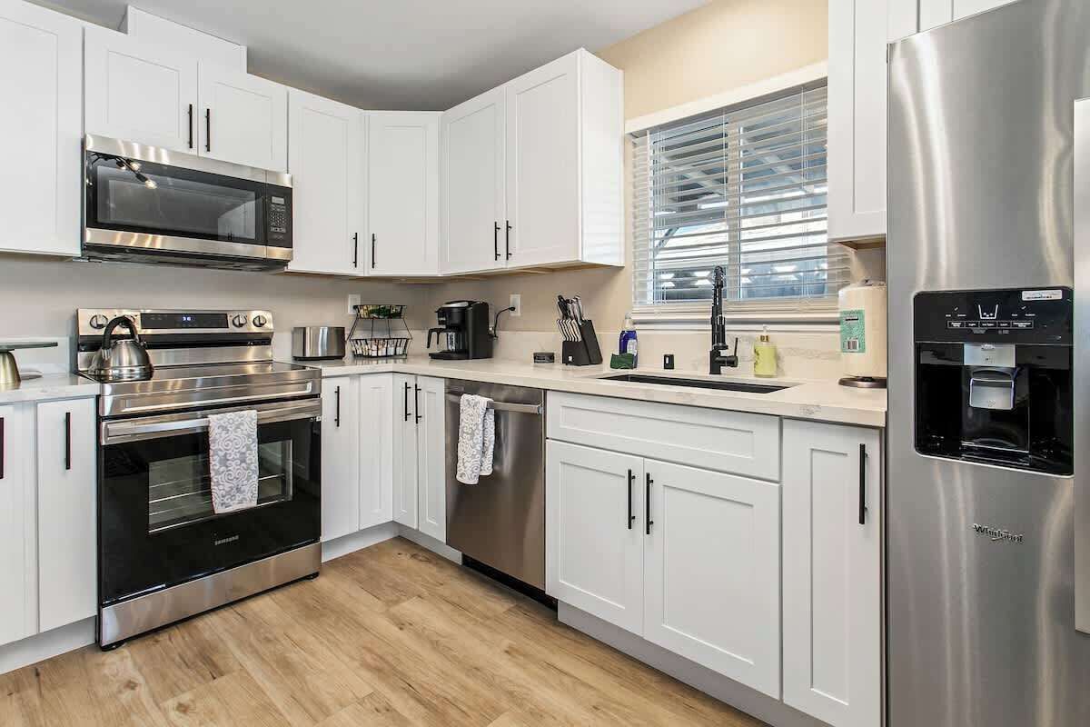 A modern kitchen with white cabinets, stainless steel appliances, and a window with blinds.