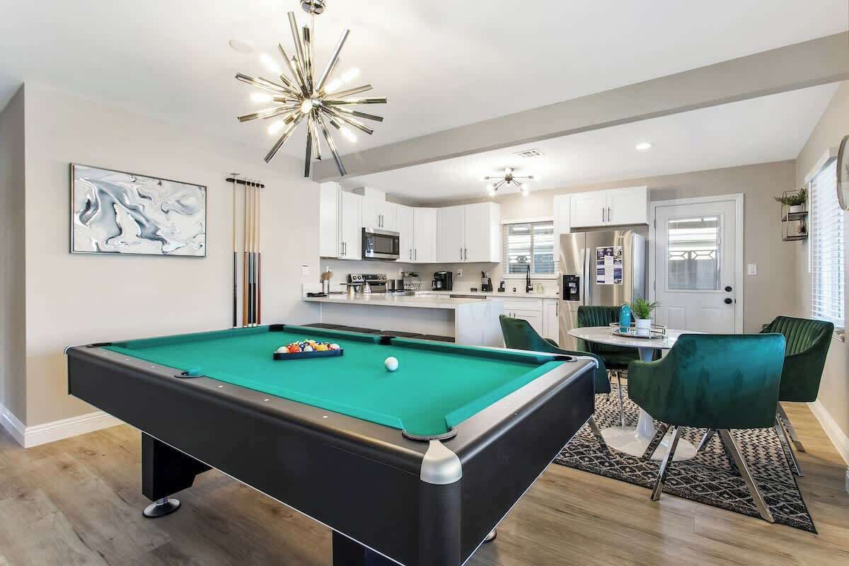 A modern living room with a pool table, a dining area with green chairs, and a kitchen in the background.