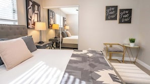 Moderni hotellihuone, jossa on suuri sänky, yöpöytä, lamppu, tuoli, pieni pöytä, jossa on kasvi, sekä taideteoksia seinillä.