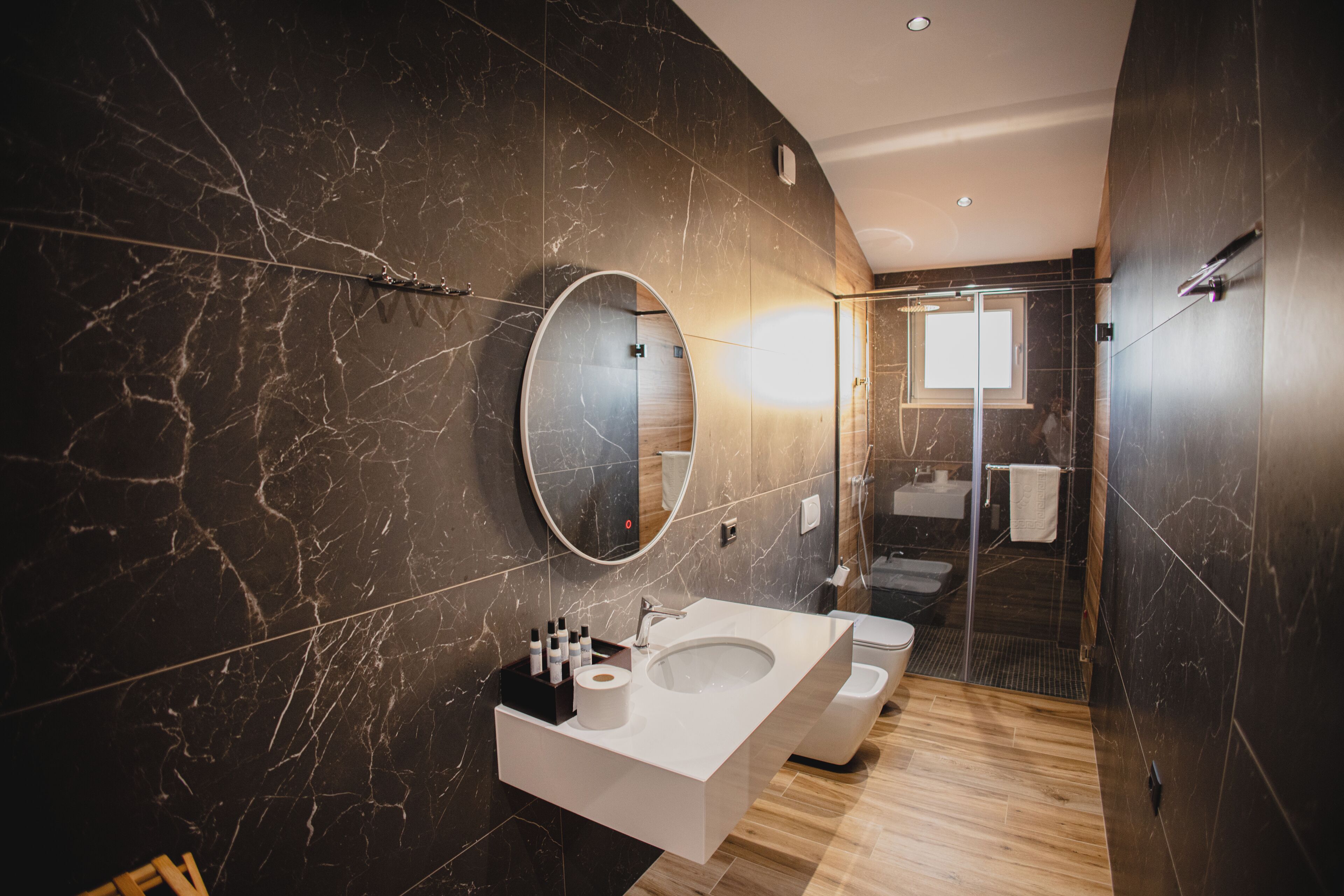 A modern bathroom with a marble wall, a large mirror, a white sink, and a shower area.
