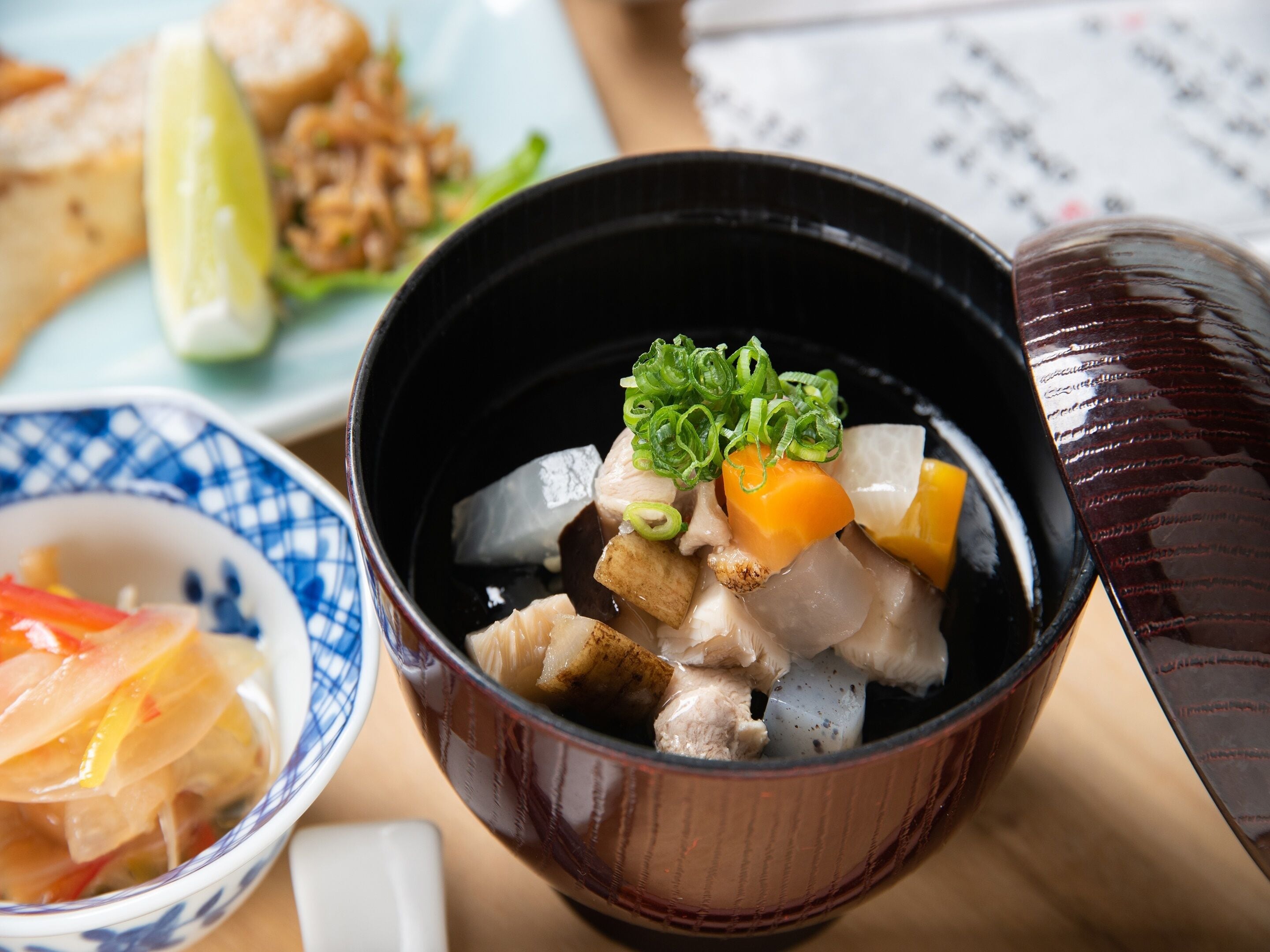 A bowl of soup with various ingredients, a plate with food, and a cup.
