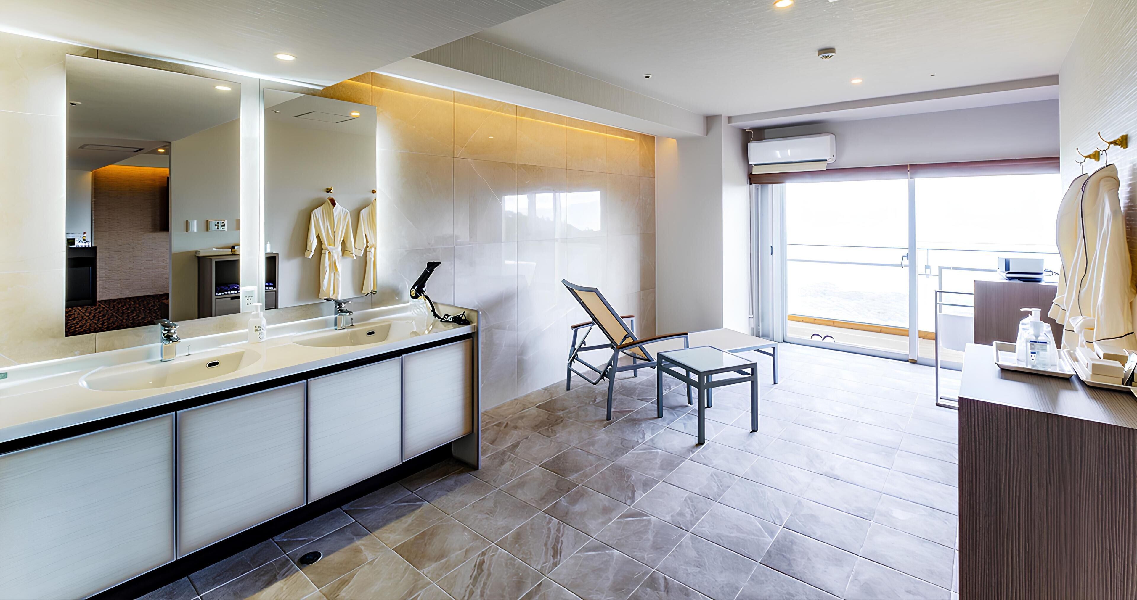 A modern bathroom with a large mirror, dual sinks, and a seating area with a chair and table.