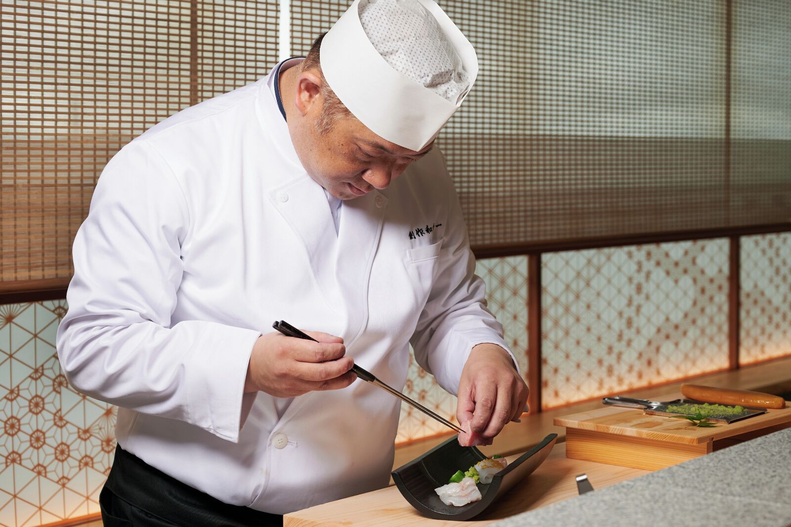 Un chef con uniforme blanco preparando comida en una cocina con estética tradicional japonesa, que incluye una mesa de madera y un revestimiento mural decorado.