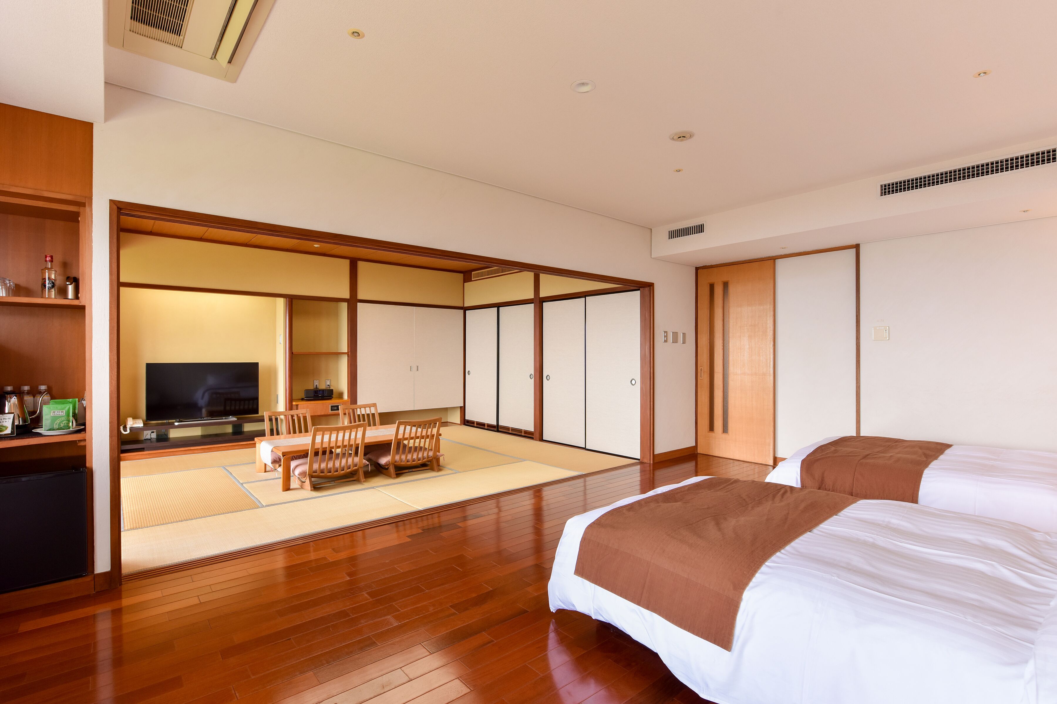 Habitación de hotel con piso de madera, cama con ropa de cama blanca y marrón, televisor, una pequeña mesa con sillas y una puerta corrediza que da a una habitación tradicional japonesa.