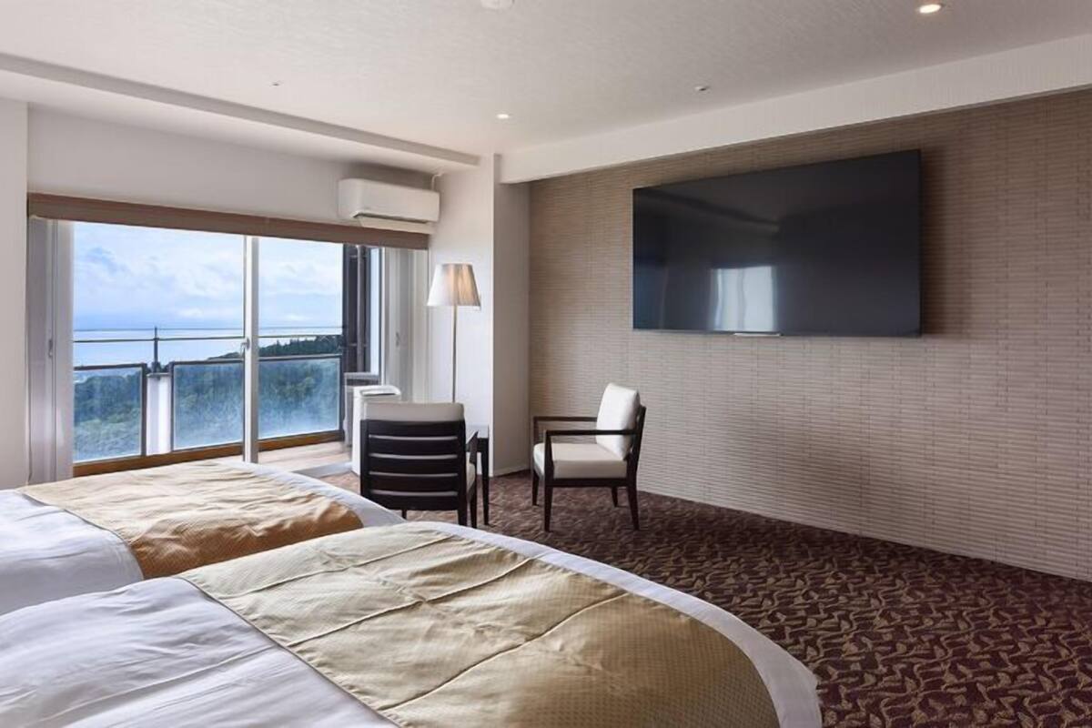 A hotel room with a large bed, a desk, a chair, a TV, and a balcony with a view.