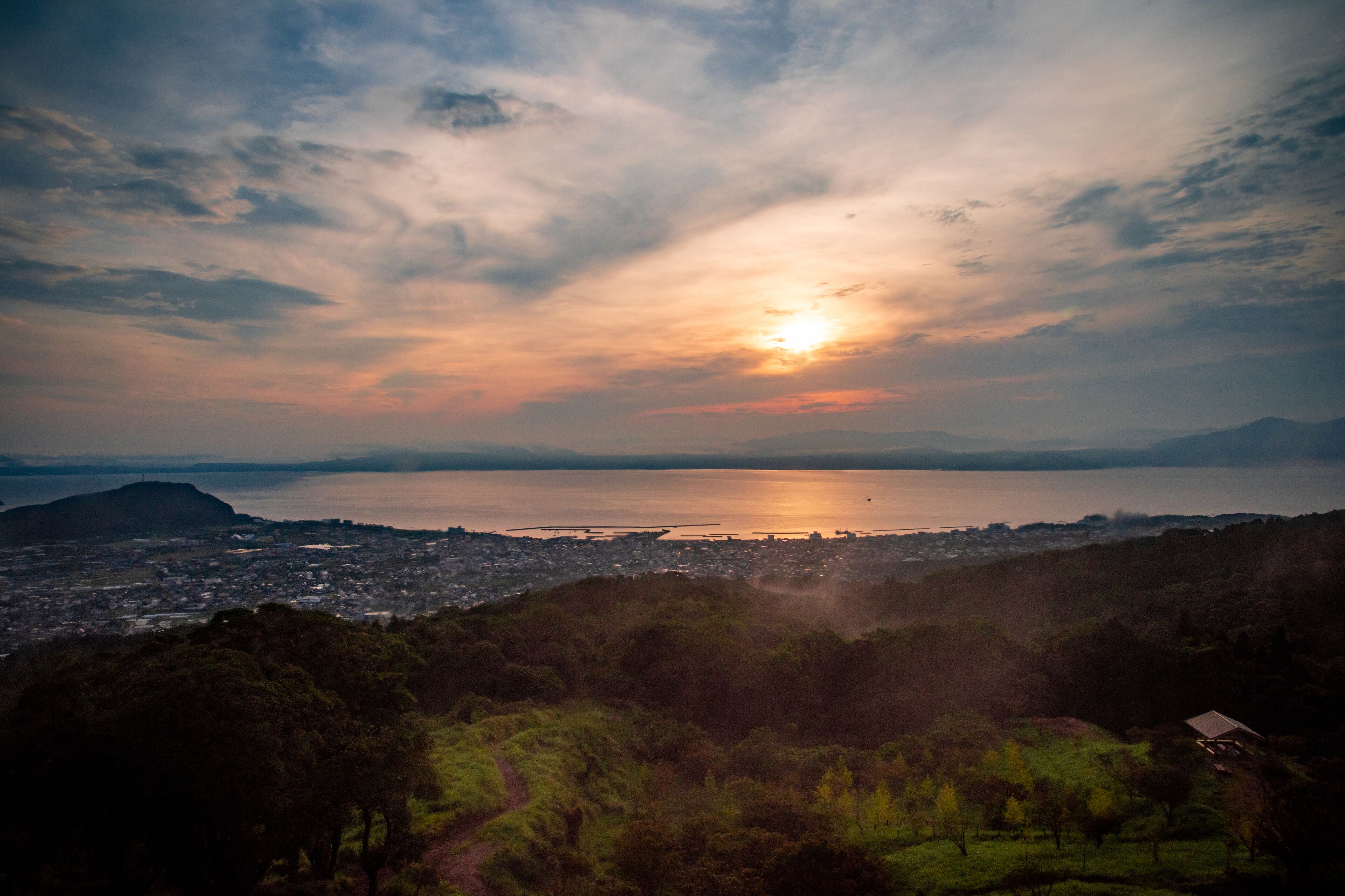 A panoramic view of a coastal city at sunset with a clear sky, a large body of water, and a distant mountain range.