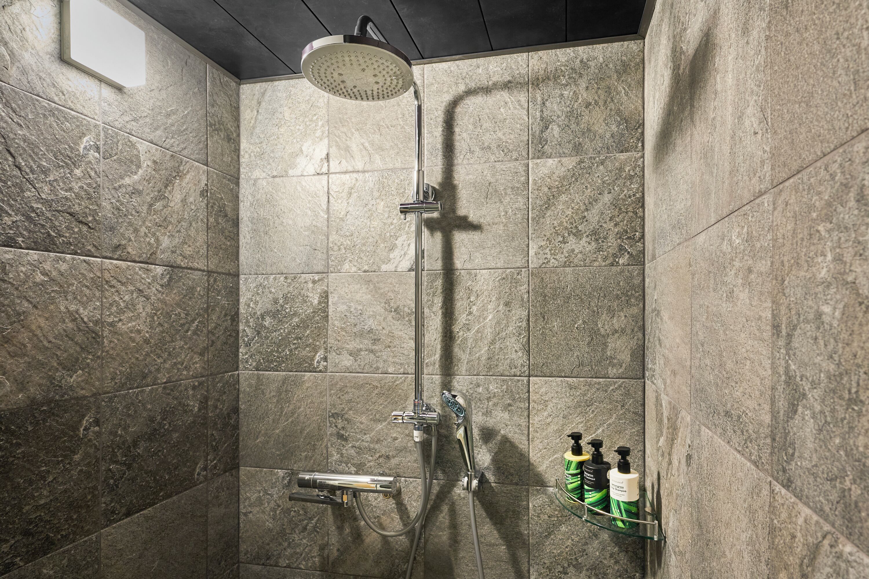 A shower with a handheld showerhead, a wall-mounted faucet, and a handheld showerhead. There is a soap dispenser on the wall.