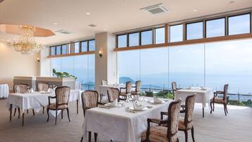 A dining area with tables set for service, large windows offering a view of the sea, and a chandelier hanging from the ceiling.