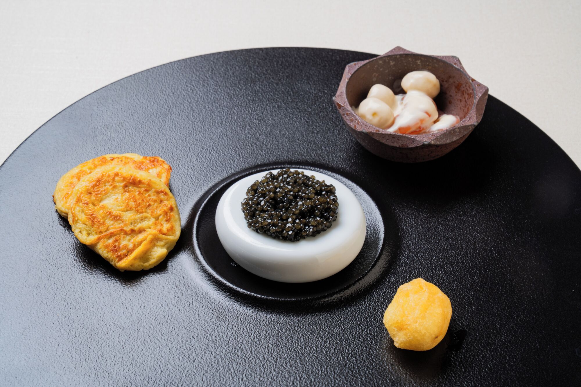 A black plate with three items: a round pastry, a bowl of caviar, and a small yellow item.