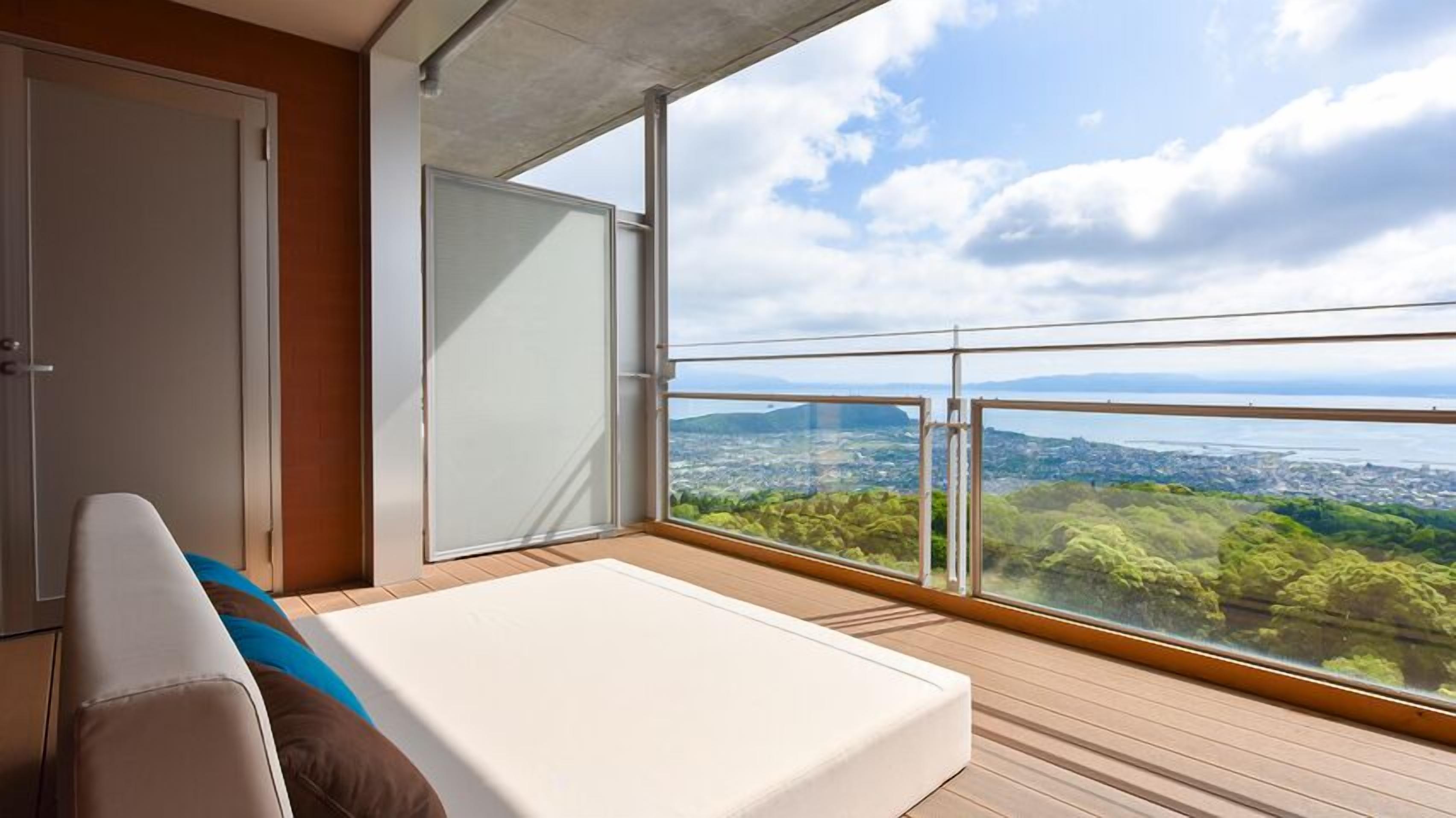 A balcony with a bed, a sliding door, and a view of a city and water.