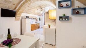 A room with a stone archway, a dining table with a bottle and glasses, a kitchen area with a microwave, and a TV mounted on the wall. - GuestHost - NEW! Apt in the heart of Monopoli! 200m from sea! (Monopoli)