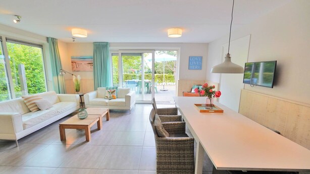 A modern living room with a white sofa, a dining table, wicker chairs, and a TV mounted on the wall.