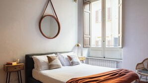 A neatly made bed with a brown throw blanket, a bedside table with a lamp, and a window with a view of buildings. - Family Suite 4 bedrooms with Terrace (Roma)