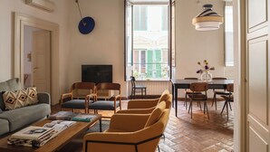 A living room with a sofa, coffee table, and chairs, a dining area with a table and chairs, and a large window with shutters. - Family Suite 4 bedrooms with Terrace (Roma)