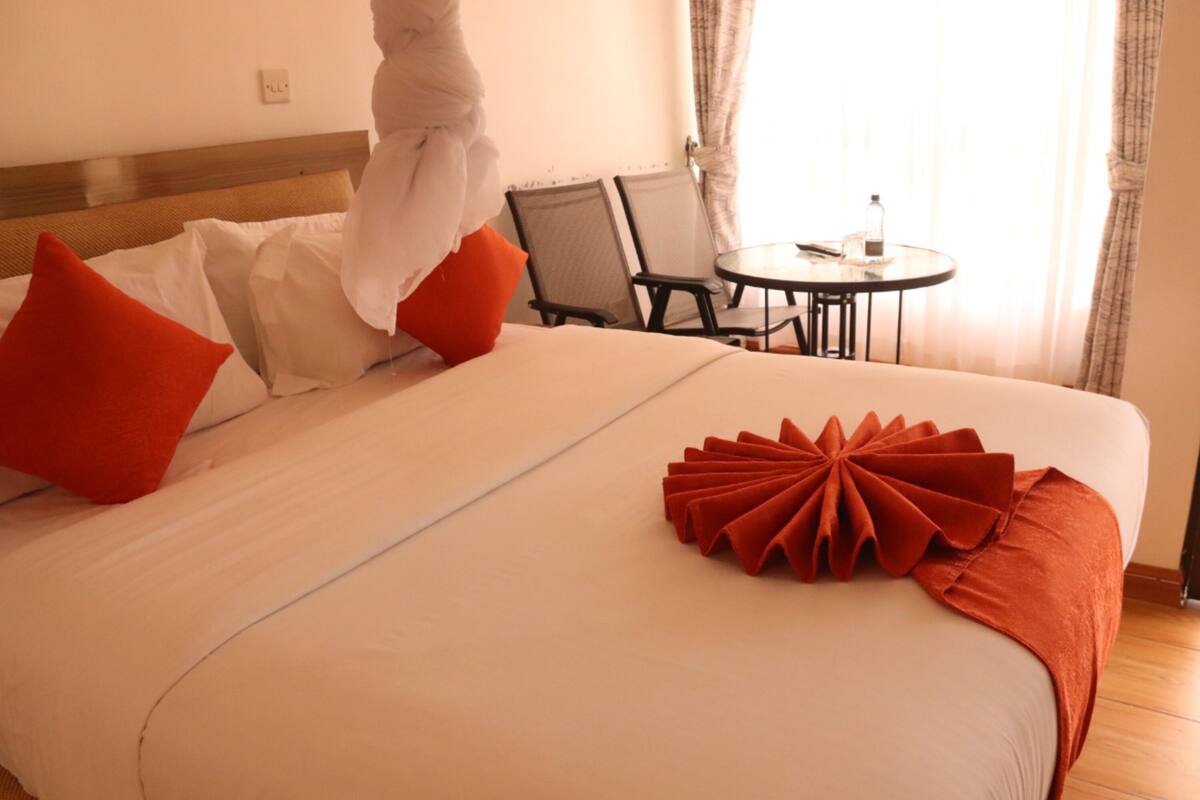 A hotel room with a neatly made bed, a red decorative pillow, and a small table with a bottle.