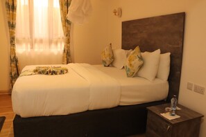 A neatly made bed with a headboard, a bedside table with a bottle and glass, and a window with sheer curtains. - Wonderland Destination Resort (Naivasha)