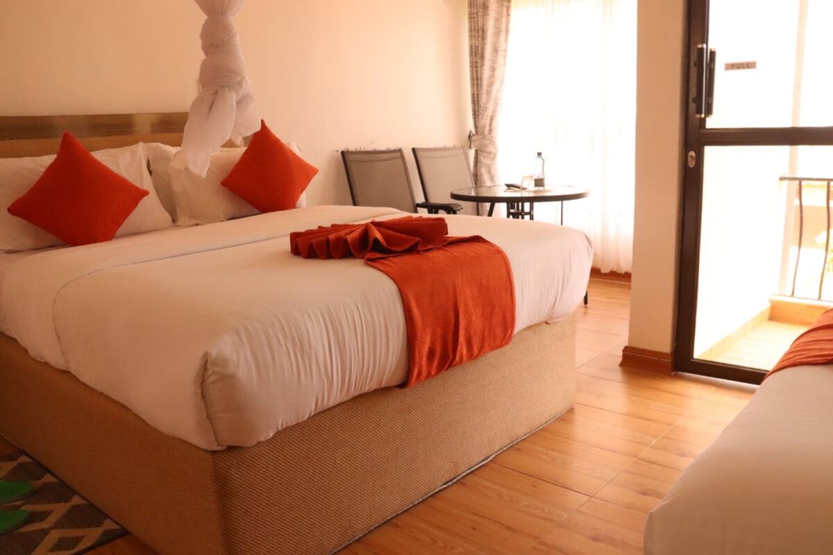 A hotel room with a large bed, two red pillows, a white canopy, a small round table, and a view through an open door.