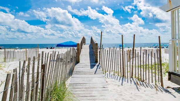 Una pasarela de madera que lleva a una playa con cielo despejado y una montaña distante.