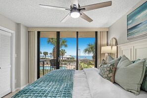 A hotel room with a bed, a ceiling fan, a view of the beach, and a pool.