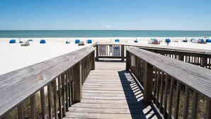 Ein Holzsteg führt zu einem Strand mit weißem Sand und blauen Stühlen.