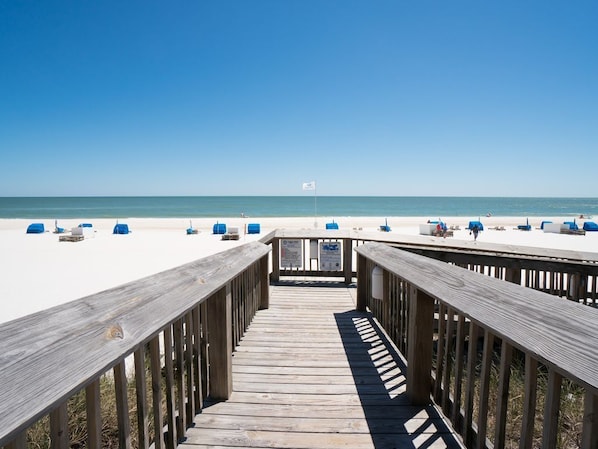 A wooden boardwalk leading to a beach with white sand and blue chairs.