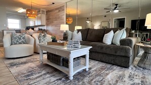 Ein modernes Wohnzimmer mit einem weißen Couchtisch, einem grauen Sofa und einem weißen Sessel. Im Hintergrund befindet sich eine Küche mit einer Holzvertäfelung und Pendelleuchten.