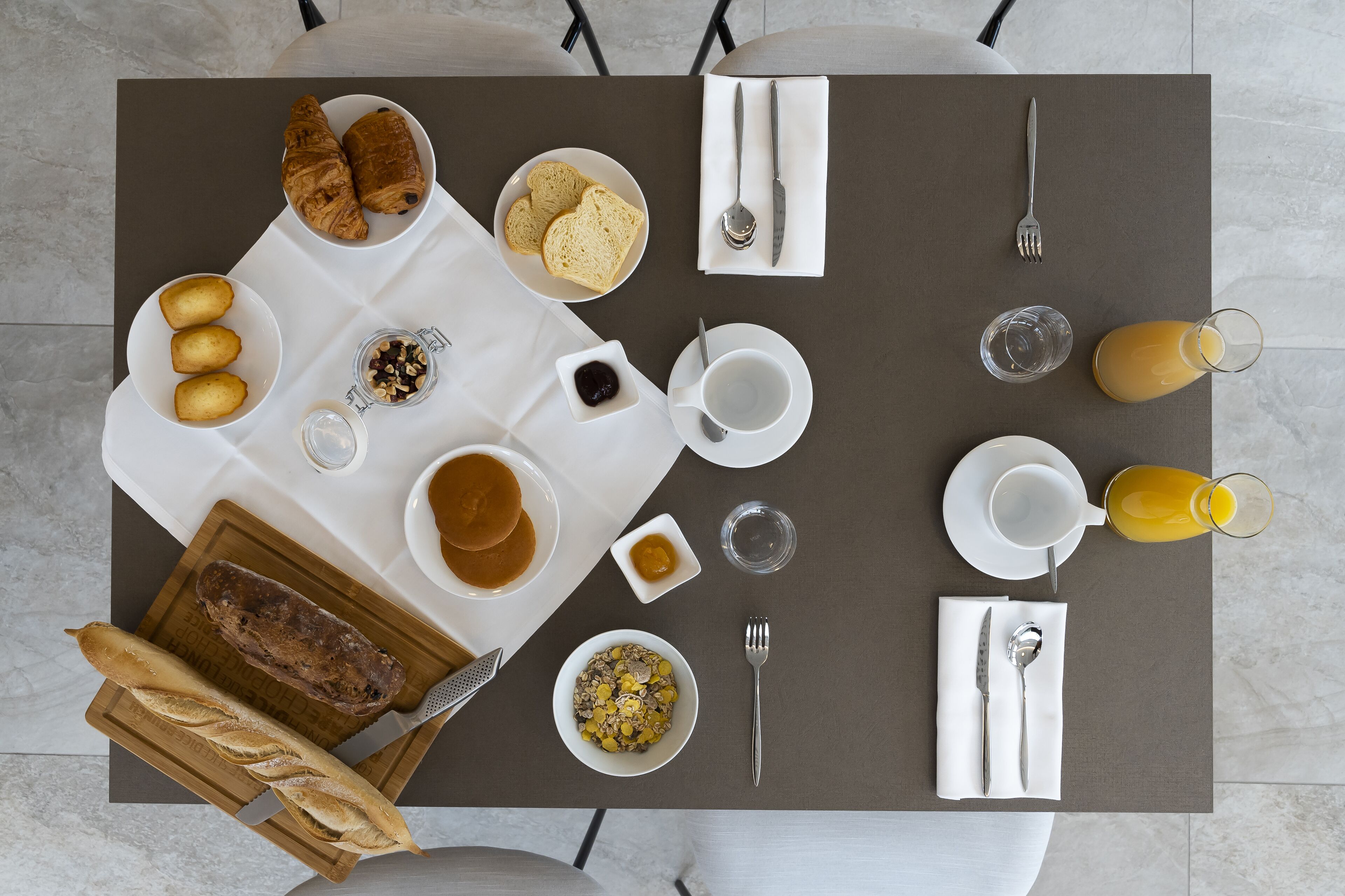 Une table dressée pour le petit-déjeuner, avec des croissants, du pain, des crêpes, des céréales et du jus de fruits.