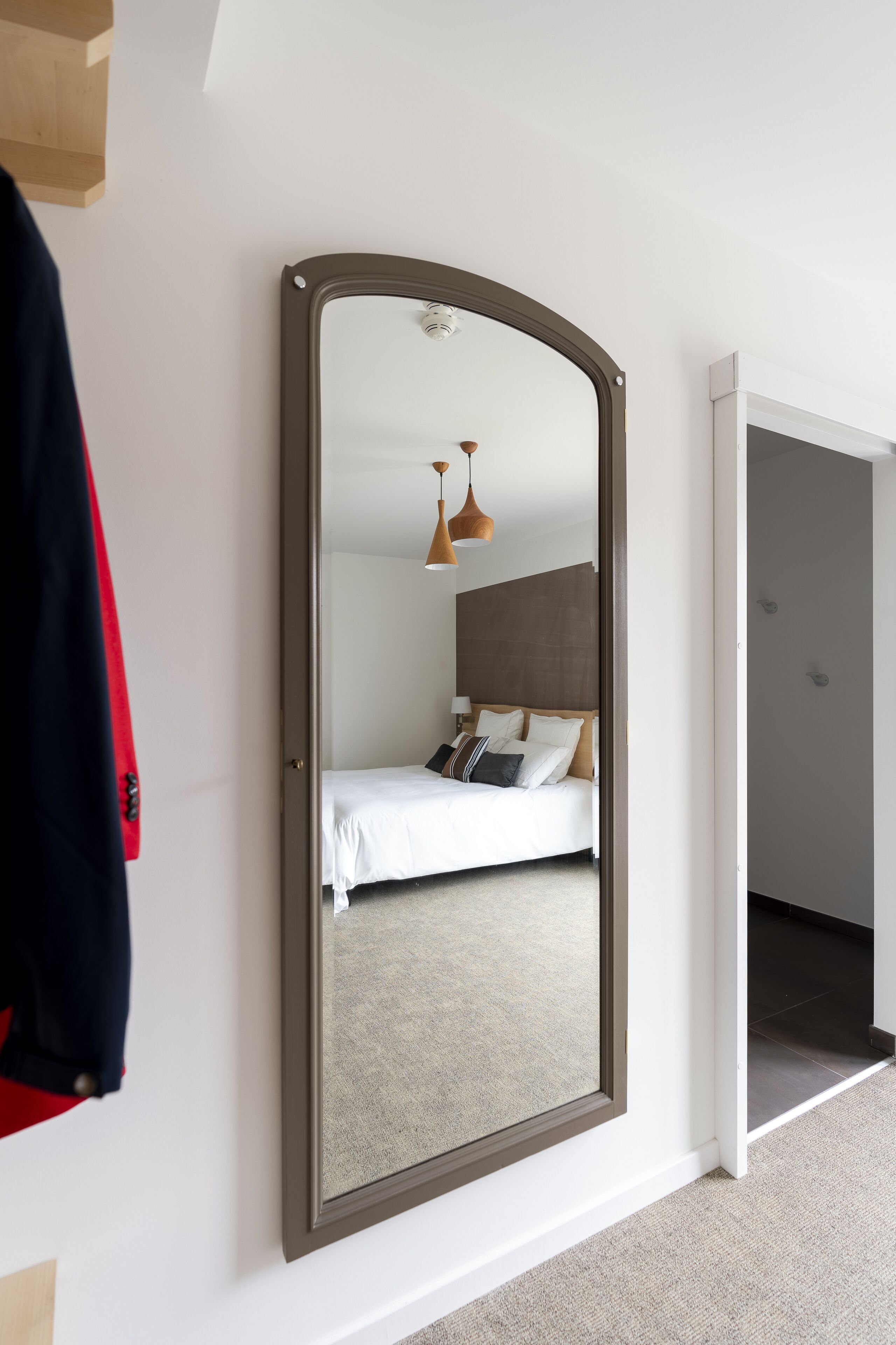 A bedroom with a large mirror, a bed with white bedding, a hanging pendant light, and a door leading to another room.
