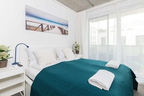 A hotel room with a bed, white nightstands, a painting of a beach, a window with curtains, and a plant.