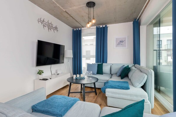A modern living room with a flat-screen TV, a sofa set, a small round table with a lamp, and a large window with curtains.