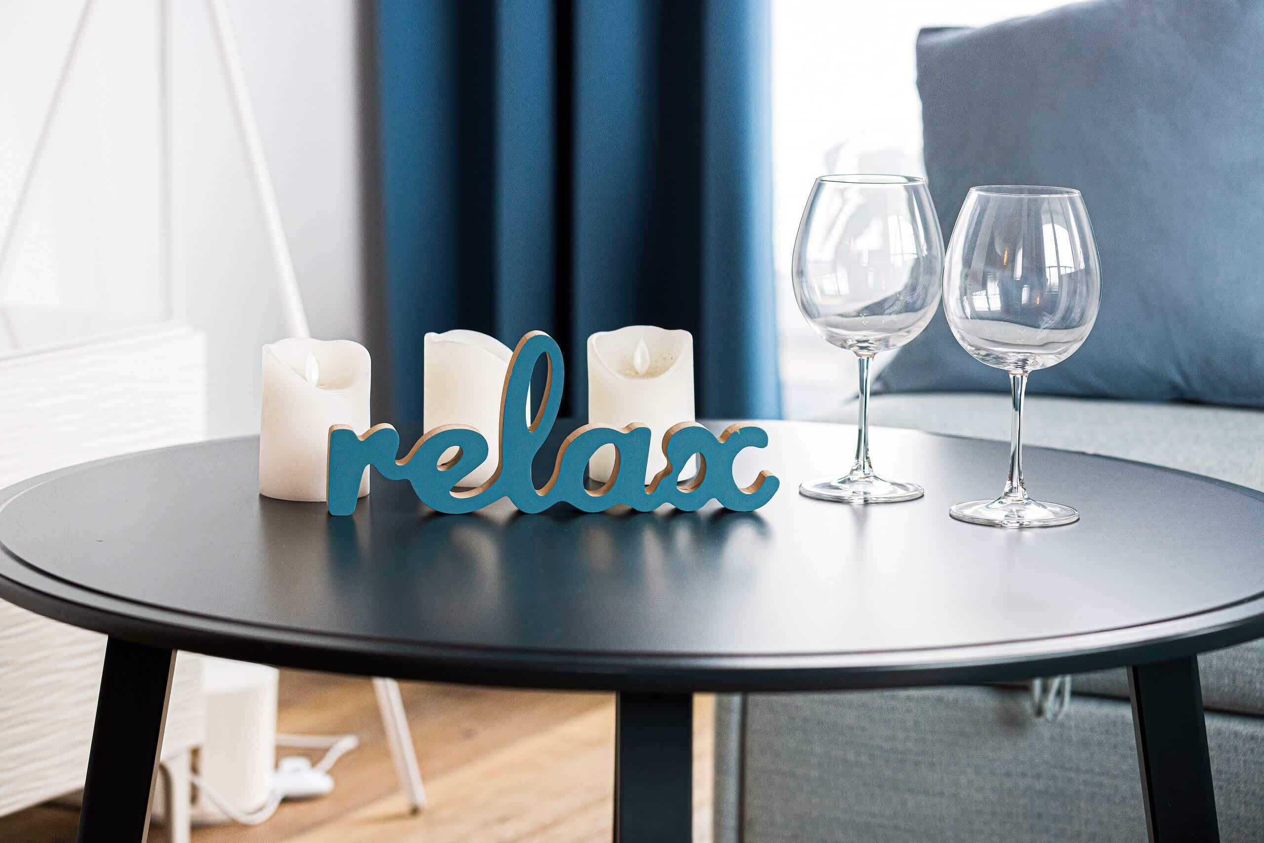 A round table with candles and two empty wine glasses, a decorative sign spelling "relax", and a blue curtain in the background.