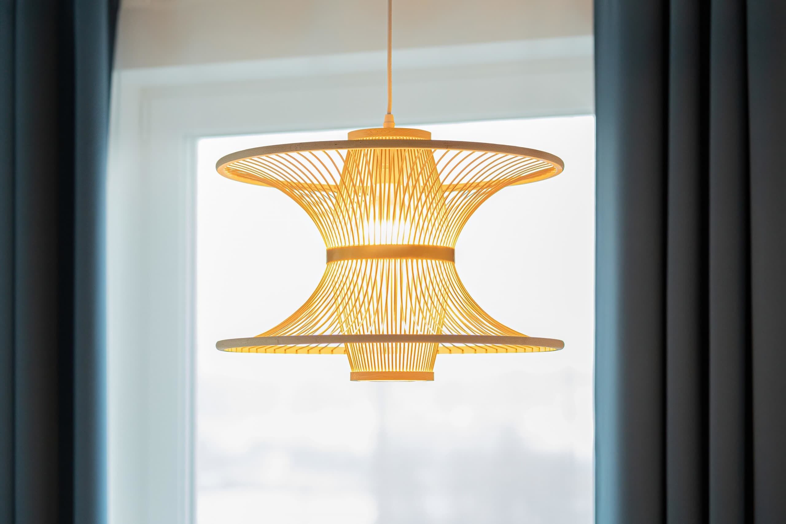 Un luminaire suspendu, de type suspension, avec un motif tissé, placé devant une fenêtre ornée de rideaux.