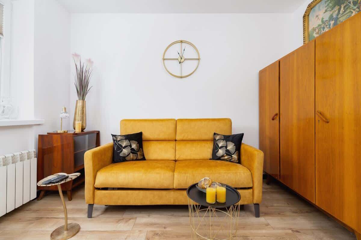 A living room with a mustard yellow sofa, a round gold clock, a wooden cabinet, and a small round table with two glasses.
