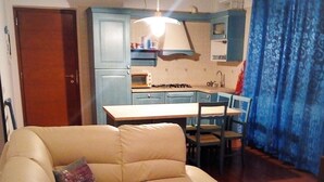 A living room with a beige sofa, a dining table with chairs, a kitchen area with blue cabinets, and a wooden door. - Downtown apartment, pets allowed (Paularo)