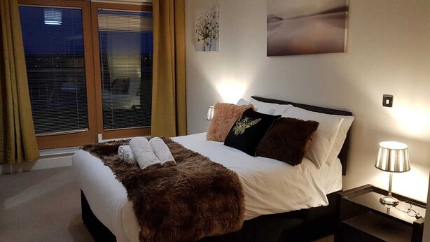 A neatly made bed with a fluffy brown throw, white pillows, and a black pillow with a design. There is a bedside lamp on a nightstand, and a window with blinds partially open.